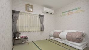 A room with a futon bed, a small shelf with toiletries, a window with curtains, and an air conditioner. - BJ Family Inn (Kyoto)