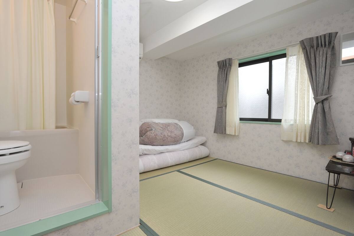 Ein traditioneller japanischer Raum mit Tatami-Böden, Futon-Bett, Toilette und einem Fenster mit Vorhängen.