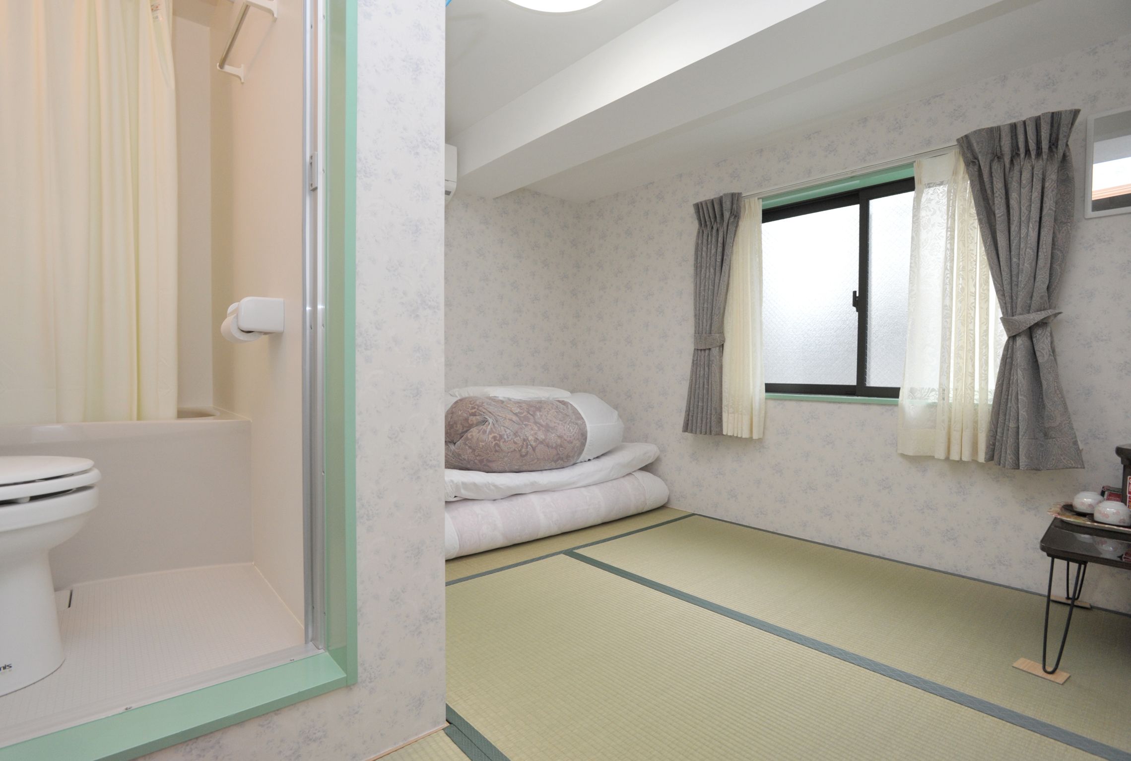 A traditional Japanese-style room with tatami flooring, a futon bed, a toilet, and a window with curtains. - BJ Family Inn (Kyoto)