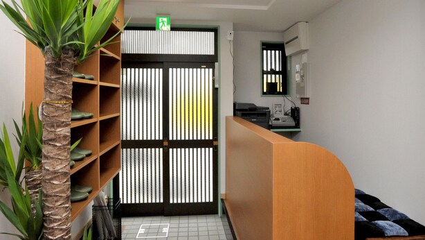 A reception area with a wooden reception desk, a palm plant, a shoe shelf, a tiled floor, and a black-framed door with a green exit sign. - BJ Family Inn (Kyoto)