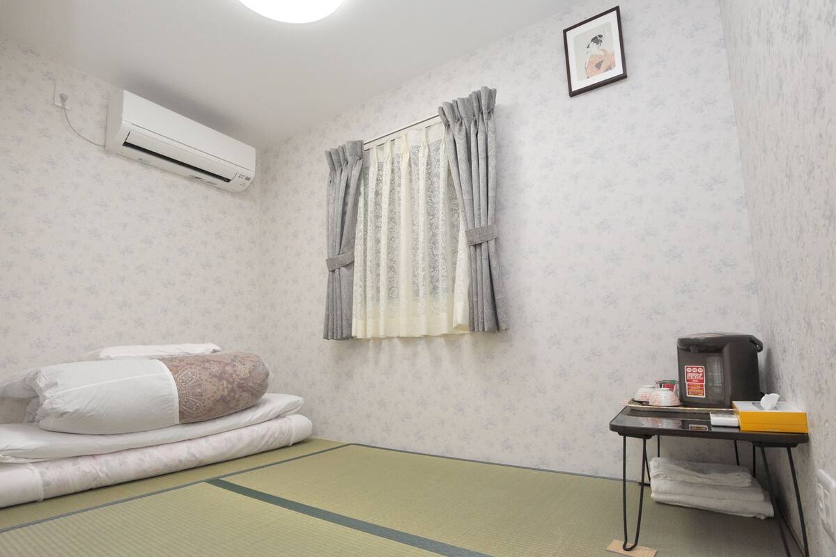 Ein Zimmer mit Tatami-Boden, einem Bett mit weißer Bettwäsche, einem kleinen Tisch mit Wasserkocher und einer Schachtel sowie einer Wand-Klimaanlage.