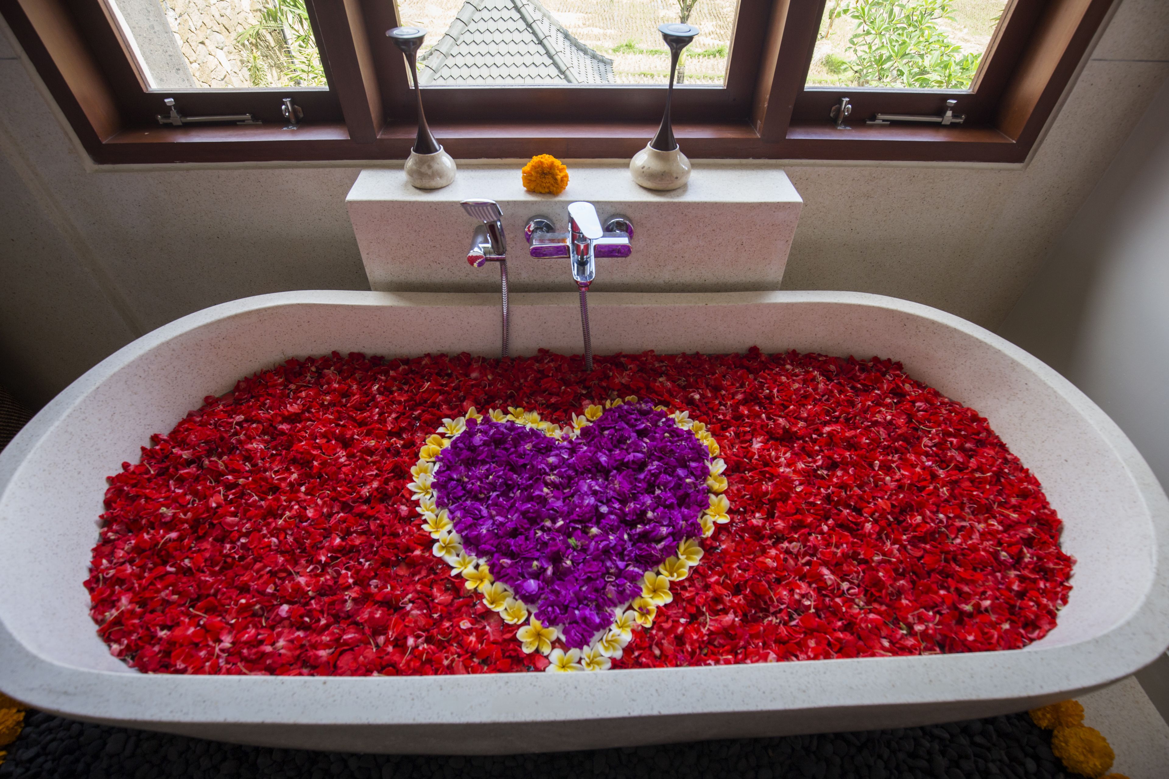 A bathtub filled with rose petals arranged in a heart shape, with a window above.