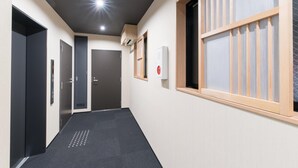 Un couloir avec un tapis noir, une fenêtre avec des stores en bois et un détecteur d’incendie fixé au mur.