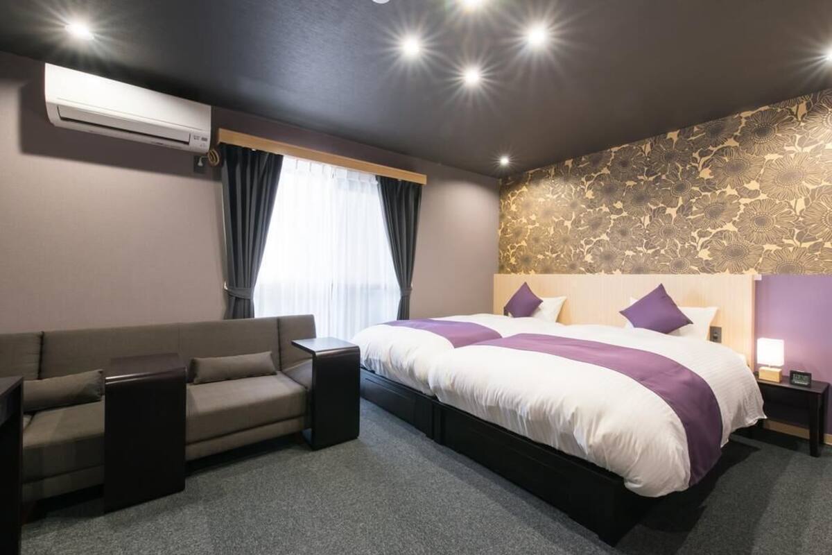Habitación de hotel con cama, sofá, mesita de noche, unidad de aire acondicionado y ventana con cortinas.