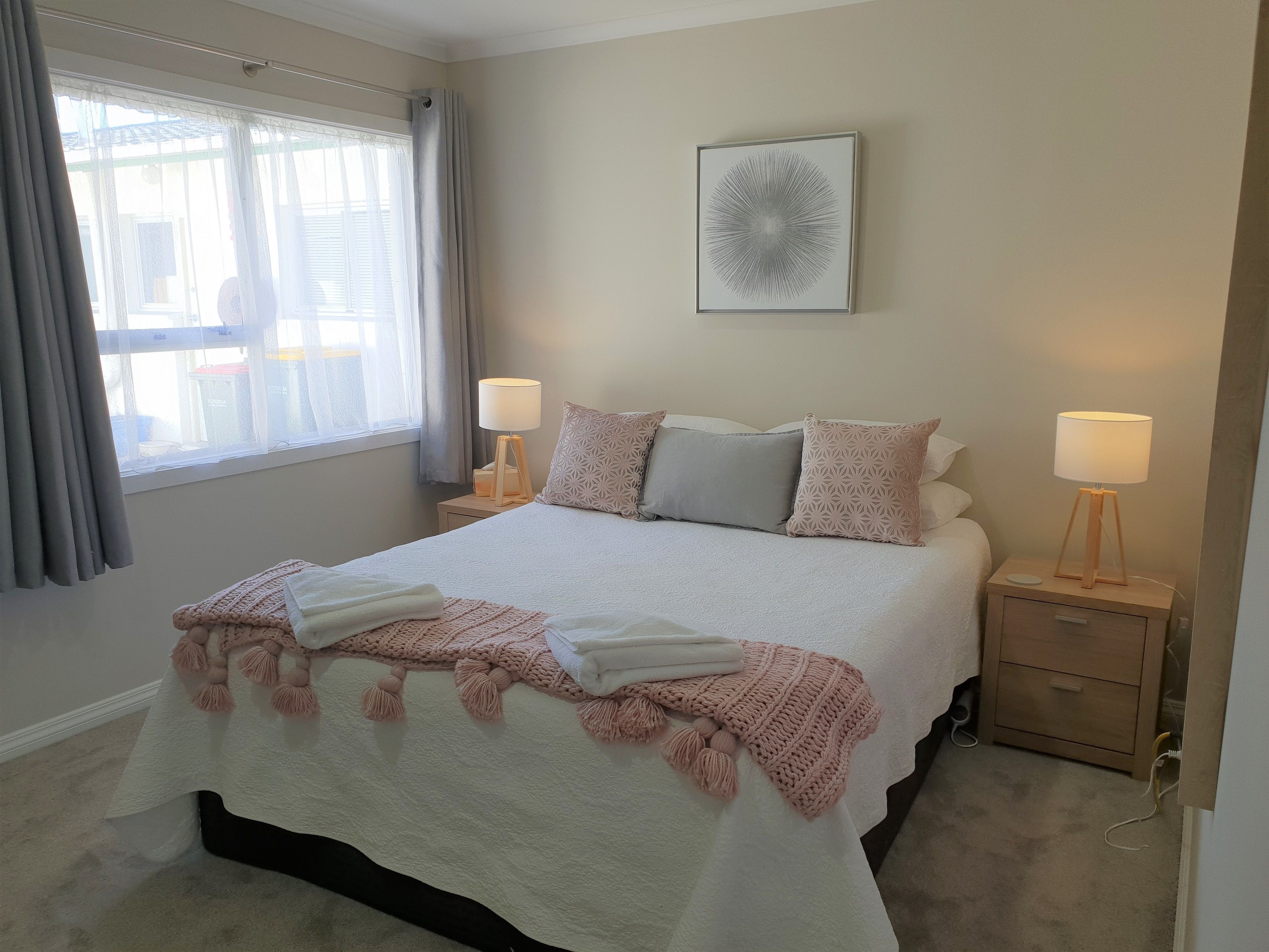 Een netjes ingerichte slaapkamer met een groot bed, twee nachtkastjes met lampen, een raam met gordijnen en een aan de muur hangend kunstwerk.