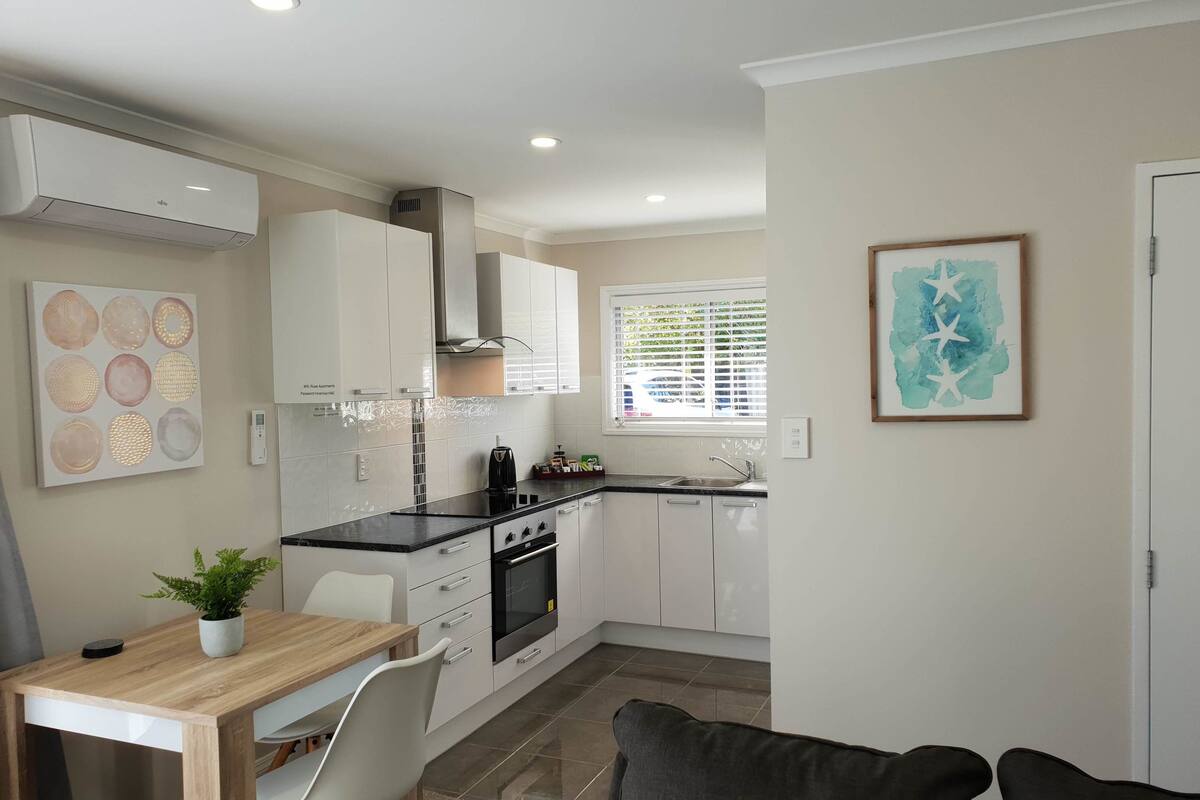 A modern kitchen with white cabinets, a black countertop, and a built-in oven. There is a dining table with chairs and a potted plant.