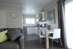 A modern kitchen with white cabinets, a dining area with a wooden table and white chairs, and a gray sofa with cushions. - Rose Apartments and Spa (Rotorua)