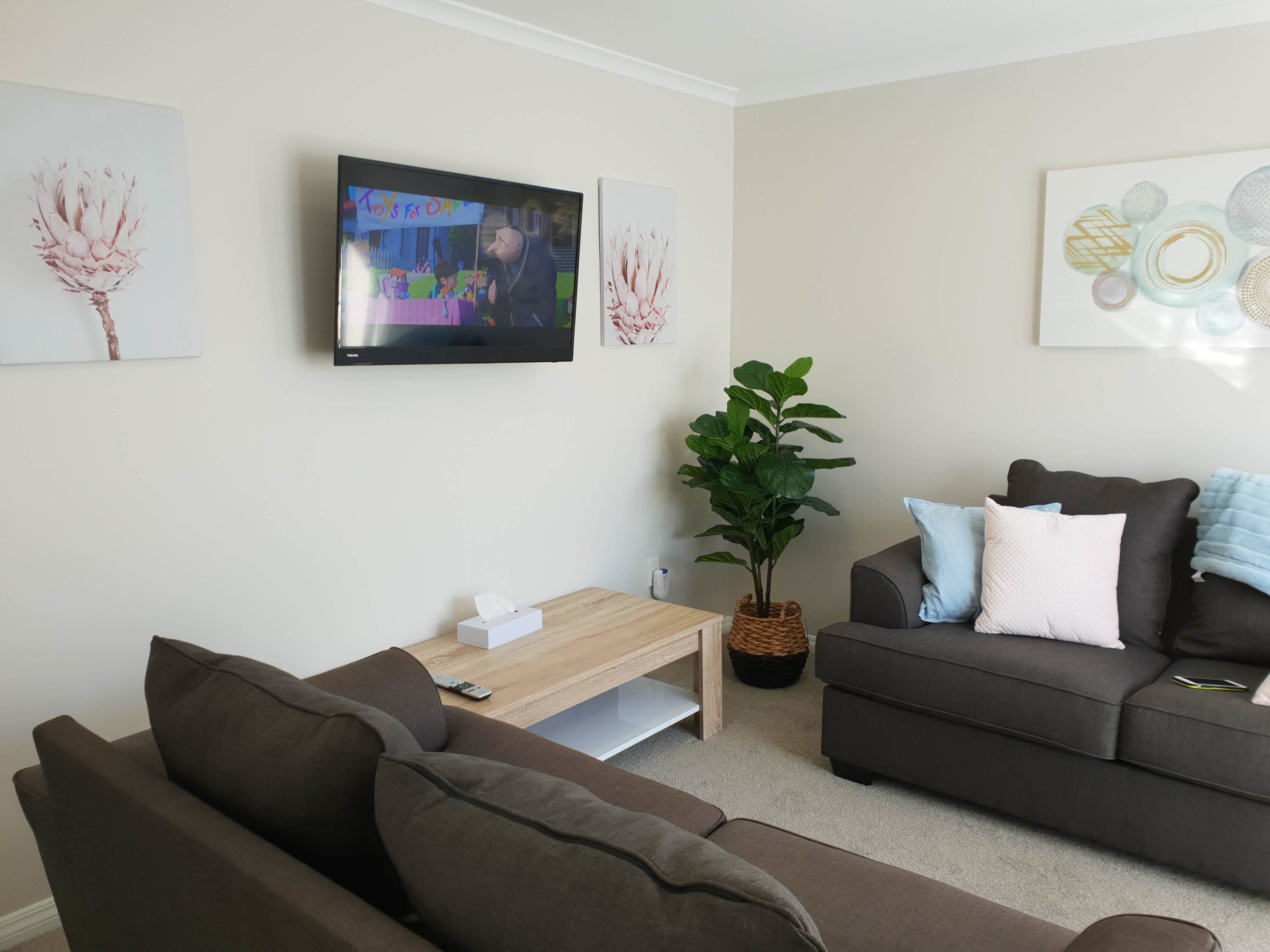 Ett vardagsrum med en mörkbrun soffa, ett träbord, en tv som är monterad på väggen och en krukväxt.