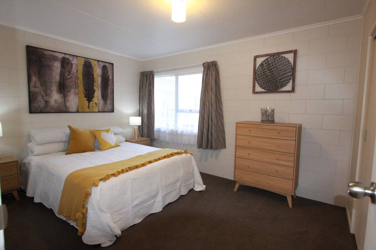 A bedroom with a large bed, a wooden dresser, a painting on the wall, and a window with curtains.