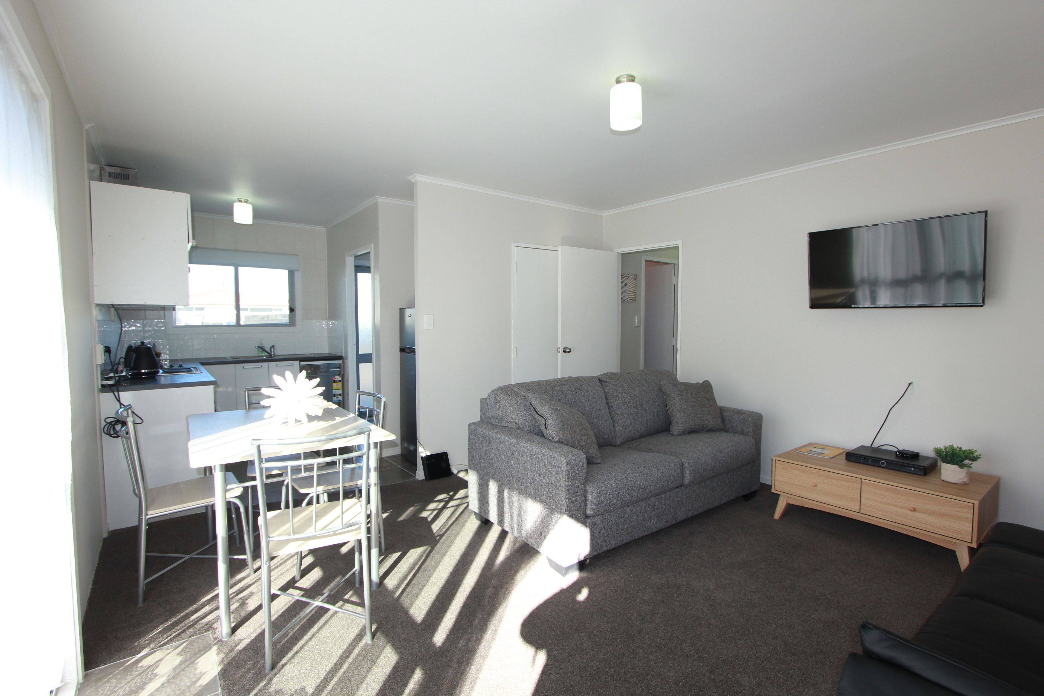 A compact living space with a kitchenette, a grey sofa, a small wooden side table, and a flat-screen TV mounted on the wall.