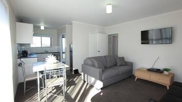 A compact living space with a kitchenette, a grey sofa, a small wooden side table, and a flat-screen TV mounted on the wall.