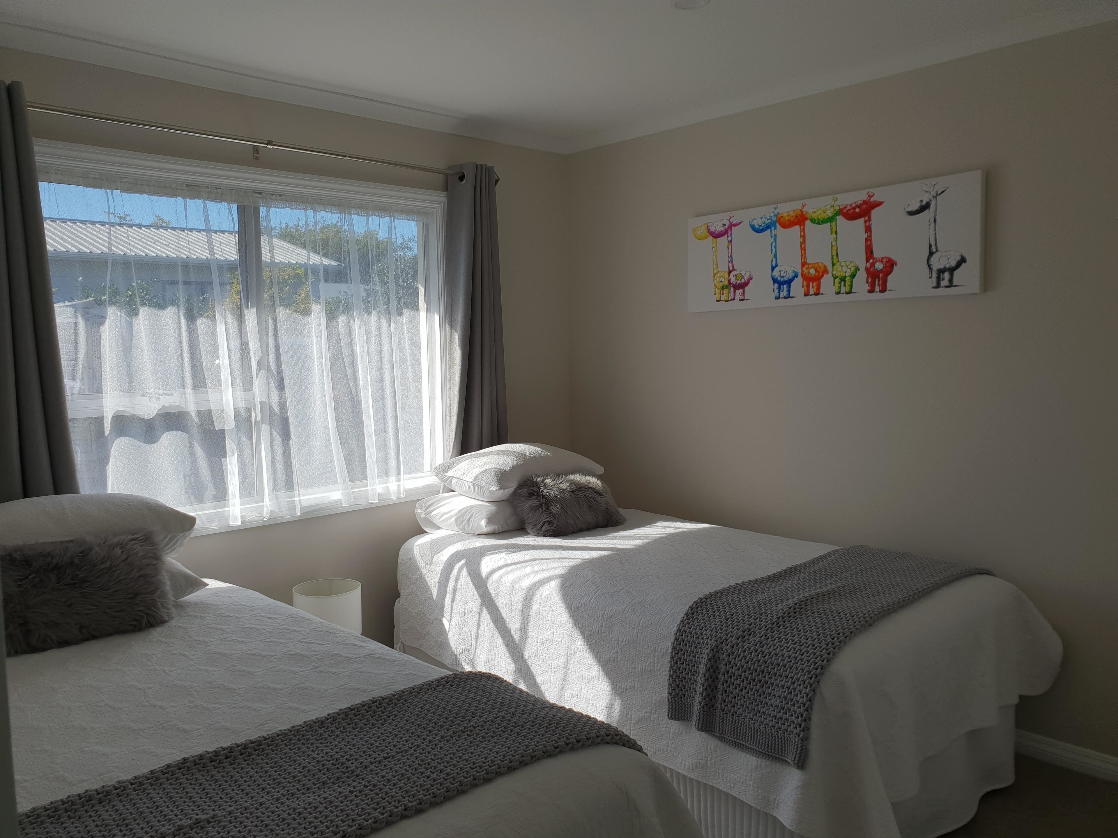 A bedroom with two beds, a window with sheer curtains, a white wall with a colorful animal painting, and a gray knitted throw blanket on one bed.