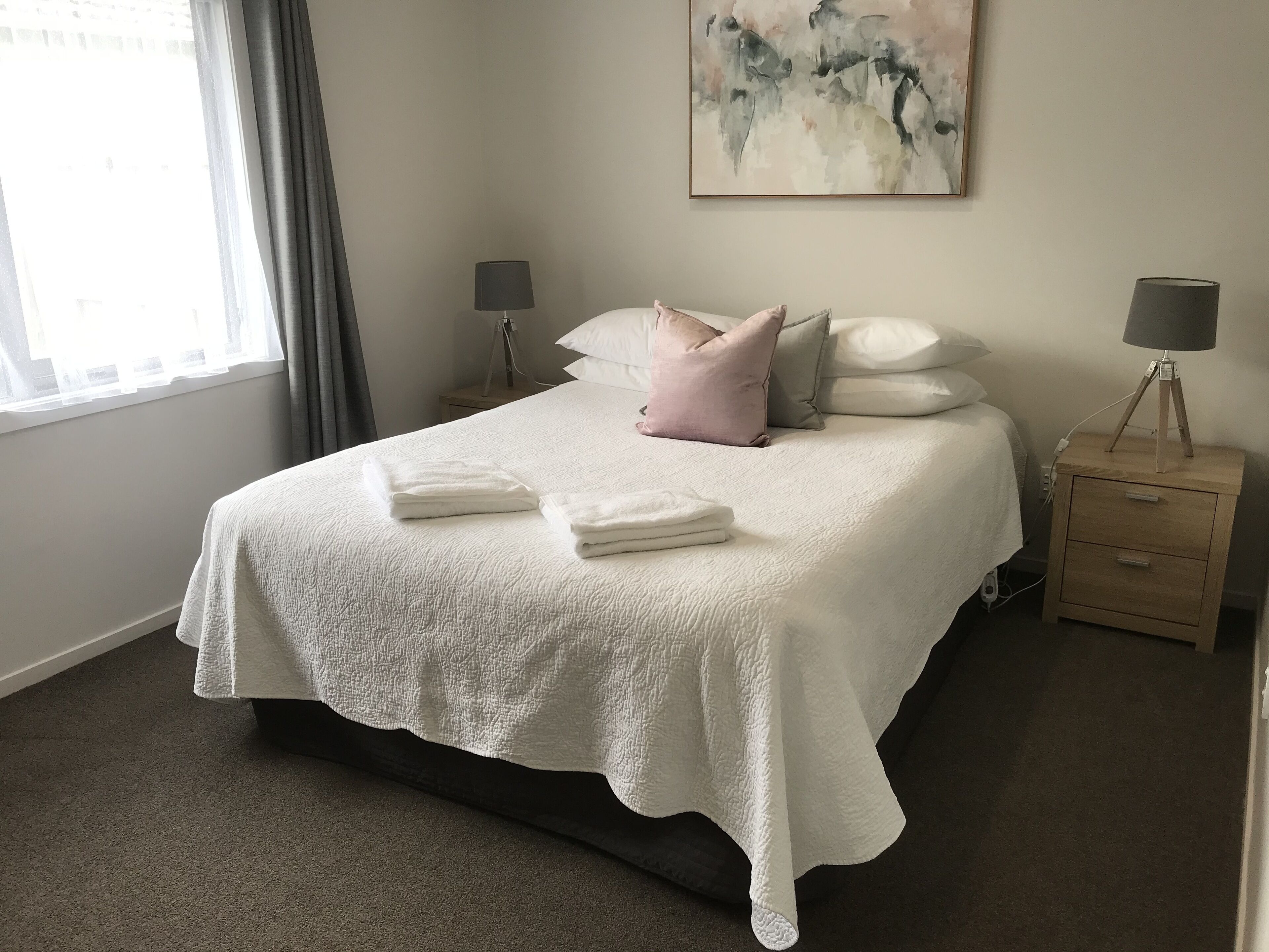 Un lit bien fait, avec du linge de lit blanc et un couvre-lit en dentelle, deux lampes de chevet, une table de chevet en bois et un tableau accroché au mur.