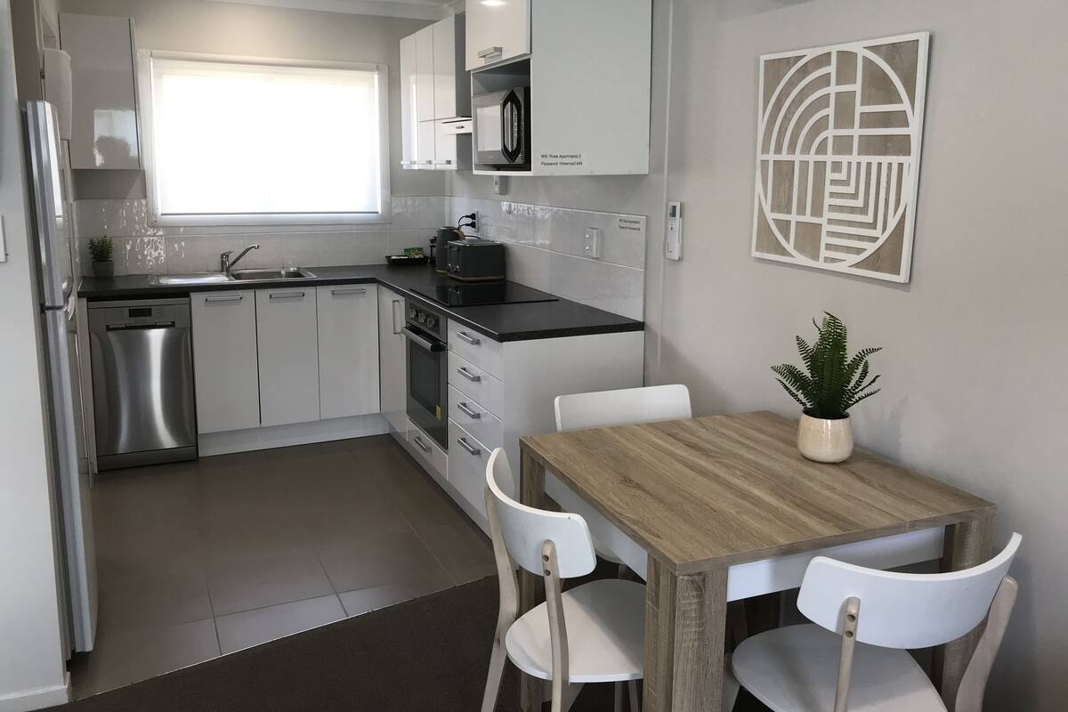 A modern kitchen with white cabinets, a stainless steel dishwasher, and a wooden dining table with two white chairs.