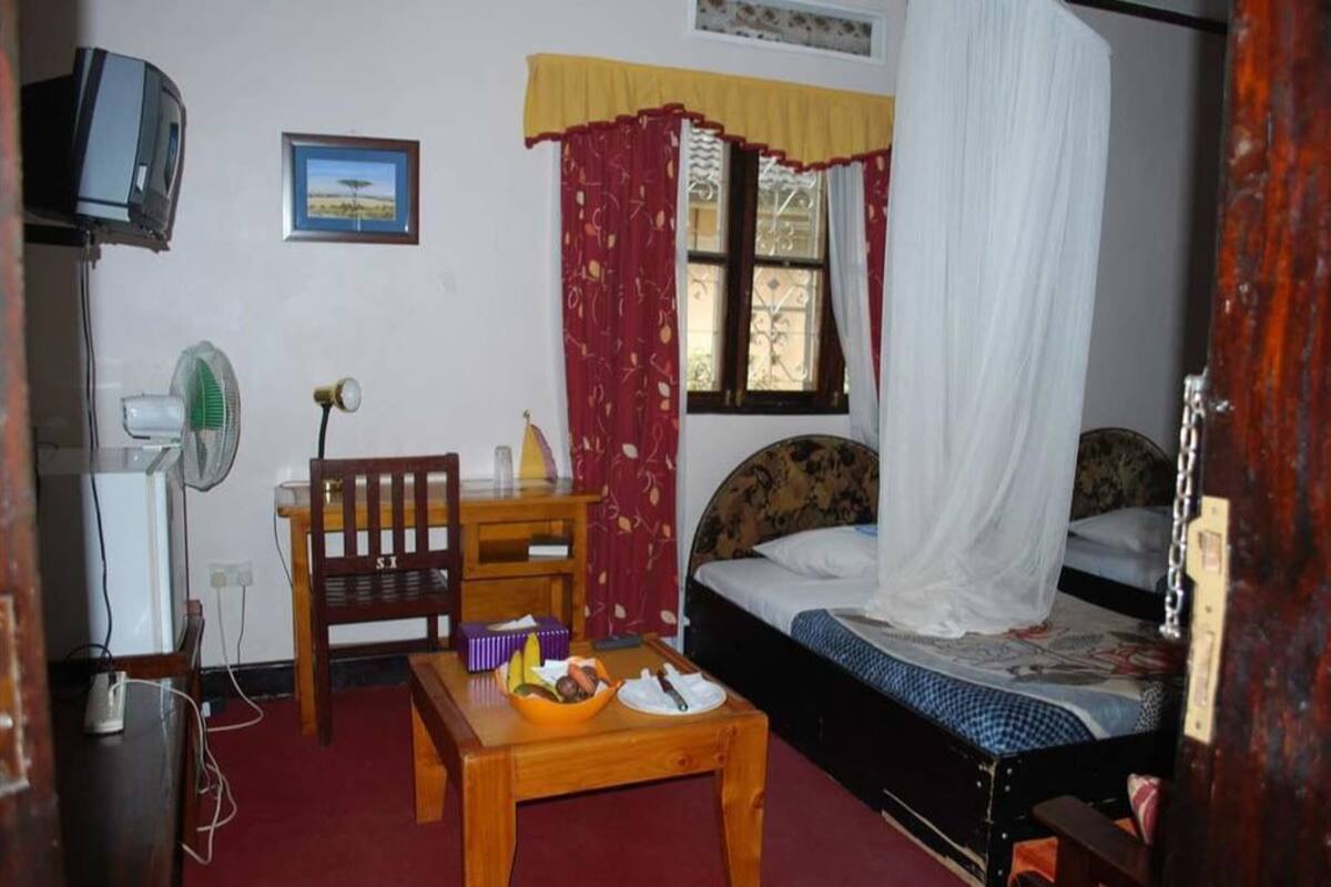 A bedroom with a bed, a desk, a chair, a bedside table, a television, and a window with curtains.