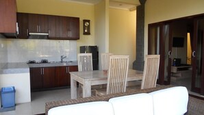 Una cocina compacta con armarios de madera oscura, estufa integrada y un lavamanos. Hay una zona de comedor con una mesa y sillas de madera, y un sofá blanco en primer plano.