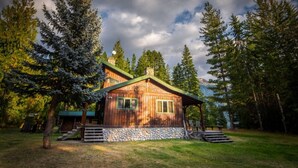 A rustic cabin with a stone foundation, wooden walls, and a green roof, surrounded by tall trees and a clear sky. - Rustic luxury in the wilderness, by the beach with lake views (Central Kootenay D)