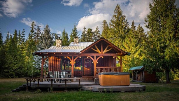 A lodge with a covered patio, hot tub, and outdoor seating area surrounded by trees. - Rustic luxury in the wilderness, by the beach with lake views (Central Kootenay D)