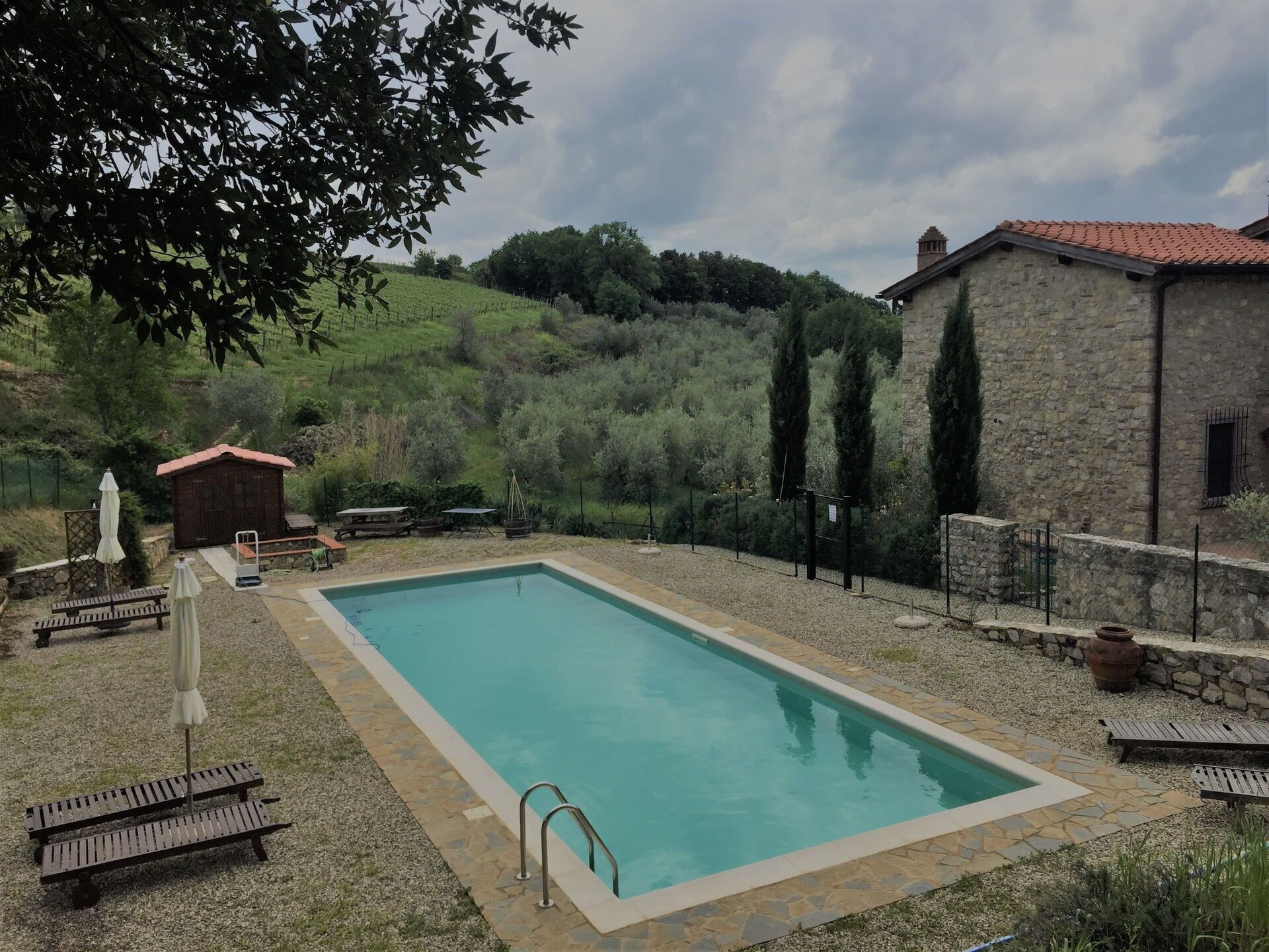 A rectangular swimming pool with a stone border, surrounded by a stone wall and a grassy area with benches and umbrellas.