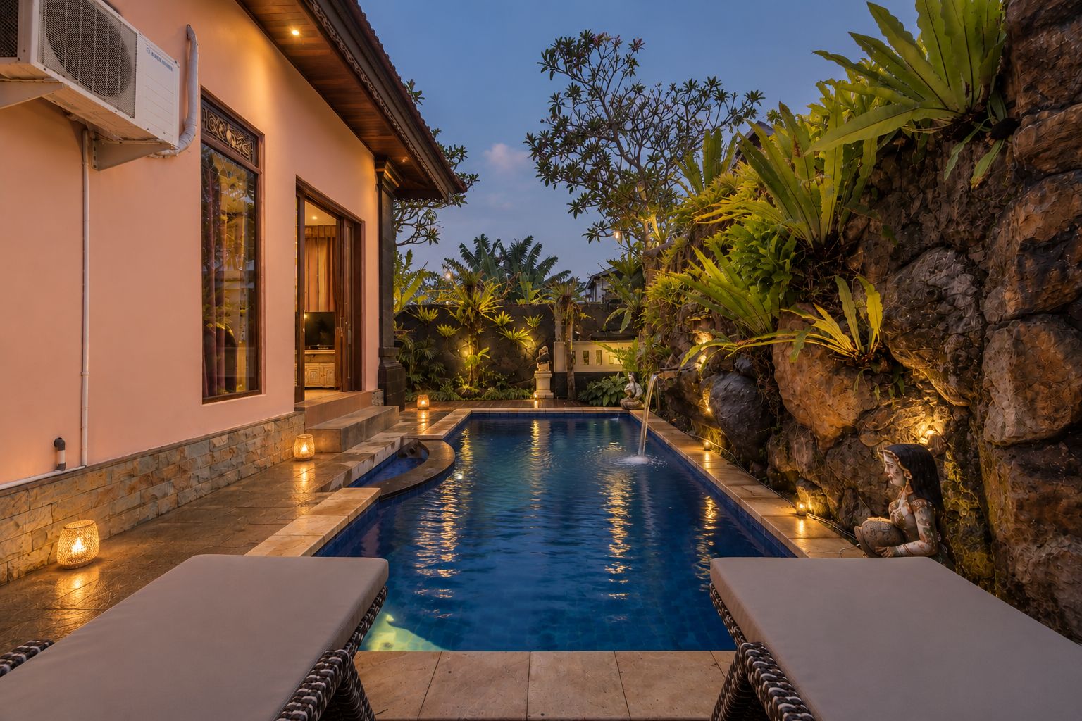 Una piscina con un muro de piedra y plantas tropicales en un lado, y un edificio con un ventanal grande y un equipo de aire acondicionado en el otro.