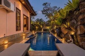 A swimming pool with a stone wall and tropical plants on one side, and a building with a large window and an air conditioning unit on the other.