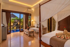 A hotel room with a four-poster bed, a sitting area with a sofa and armchairs, a small table, and a view of a swimming pool and outdoor area.