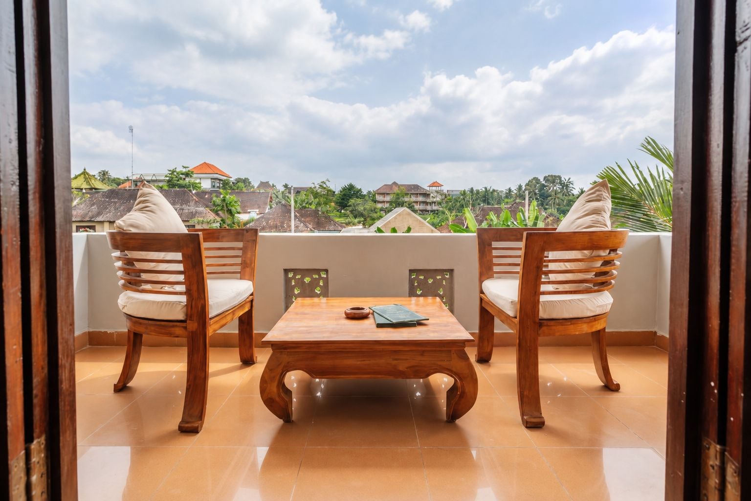 Un balcón con dos sillas de madera y una mesa, piso de baldosas y vistas a una zona residencial con casas y vegetación.