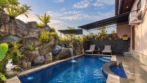 A swimming pool with a waterfall feature, surrounded by rocks and plants. There are lounge chairs and a covered seating area. - Grand Ashanti Villa (Ubud)