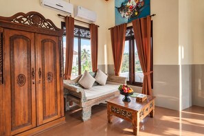 A room with a wooden cabinet, a sofa, a coffee table with fruit, and a view of palm trees through the window. - Grand Ashanti Villa (Ubud)
