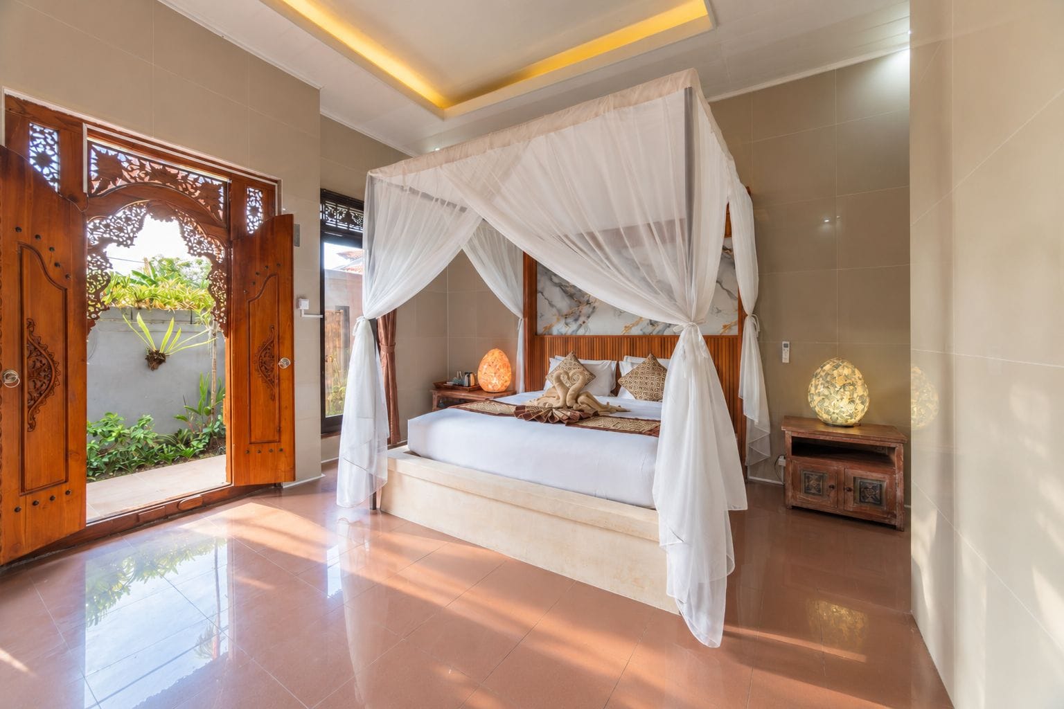 A bedroom with a four-poster bed, a nightstand with a lamp, and a view of the outdoors through a wooden door with intricate carvings.