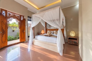 A bedroom with a four-poster bed, a nightstand with a lamp, and a view of the outdoors through a wooden door with intricate carvings. - Grand Ashanti Villa (Ubud)