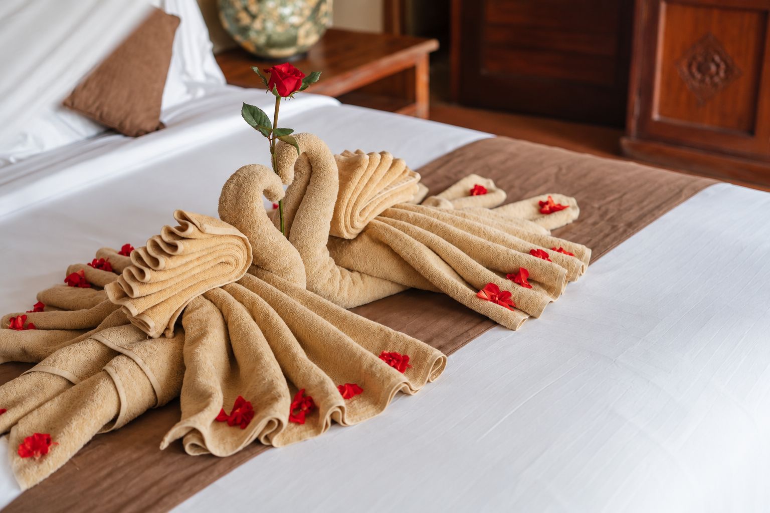 Een net opgemaakt bed met een handdoeksculptuur en een enkele rode roos.