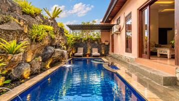 A swimming pool with a waterfall feature, surrounded by tropical plants and a stone wall.