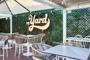An outdoor seating area with wicker chairs and tables, a sign reading 'The Yard', and a green leafy wall. - Rosehill Hotel (Granville)