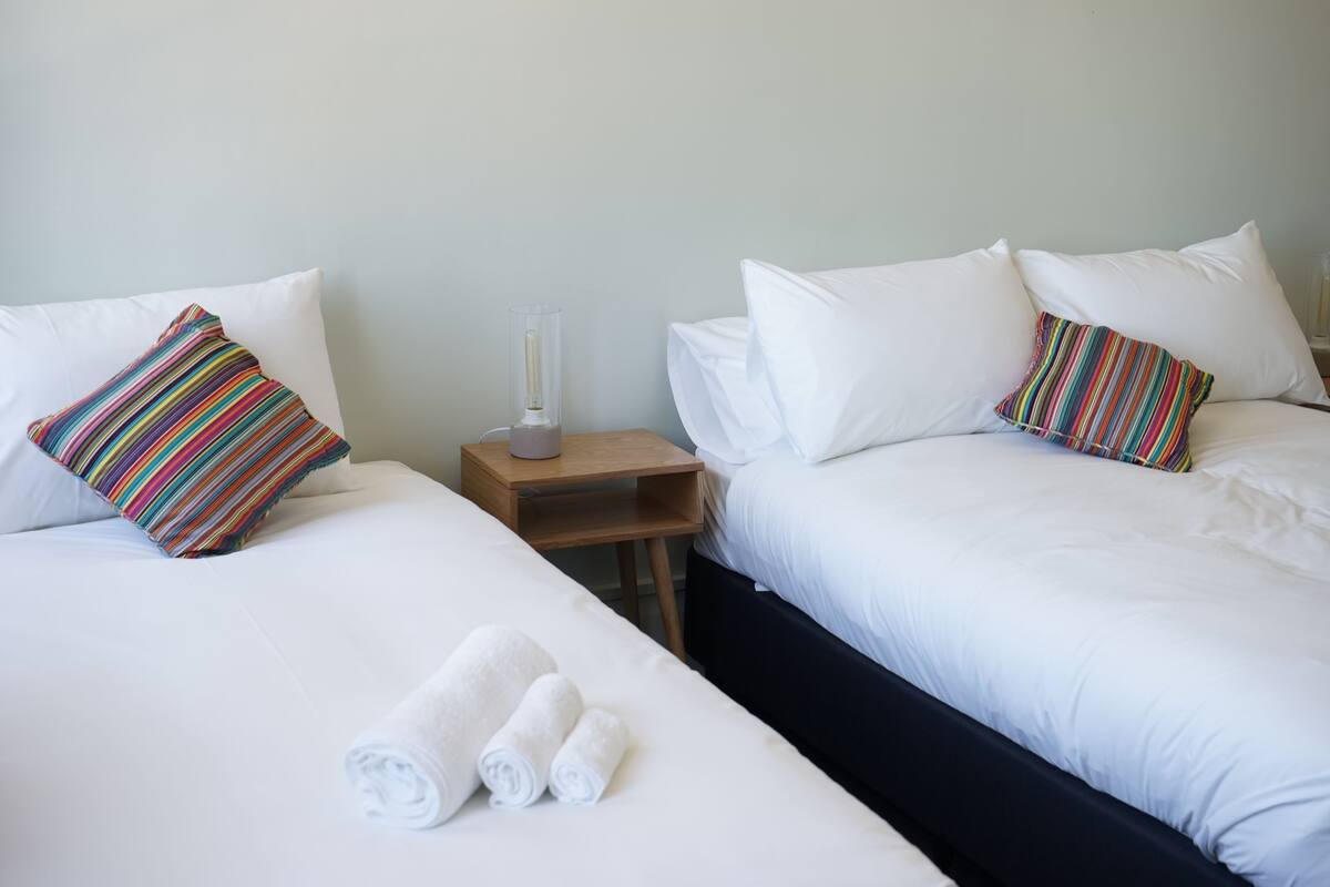 A hotel room with two beds, white bedding, striped pillows, a nightstand with a glass container, and rolled towels on the floor.