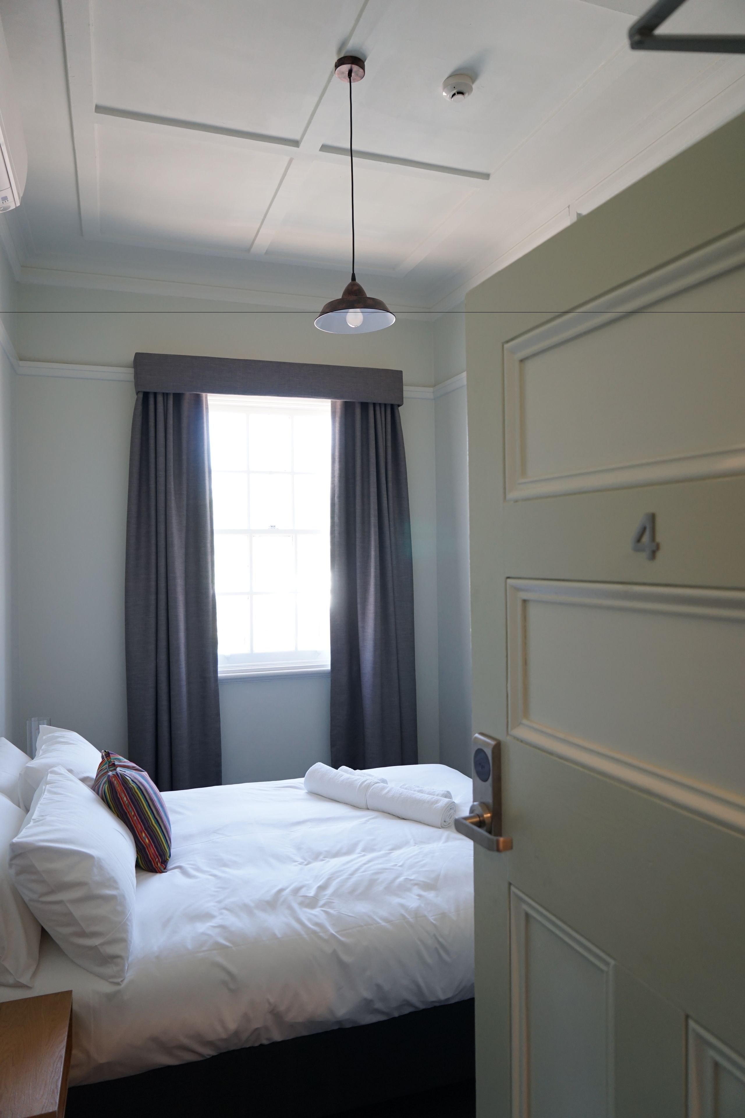 Habitación de hotel con cama, ventana con cortinas, puerta con número y una lámpara en el techo.