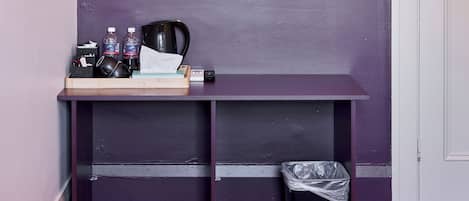 Una habitación pequeña con una pared morada, un banco de madera con un espejo, un perchero, un cesto de basura y una puerta.