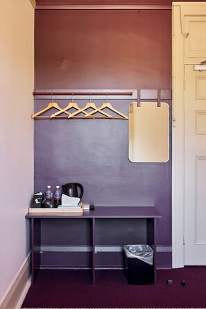 A small room with a purple wall, a wooden bench with a mirror, a coat rack, a trash bin, and a door. - Rosehill Hotel (Granville)