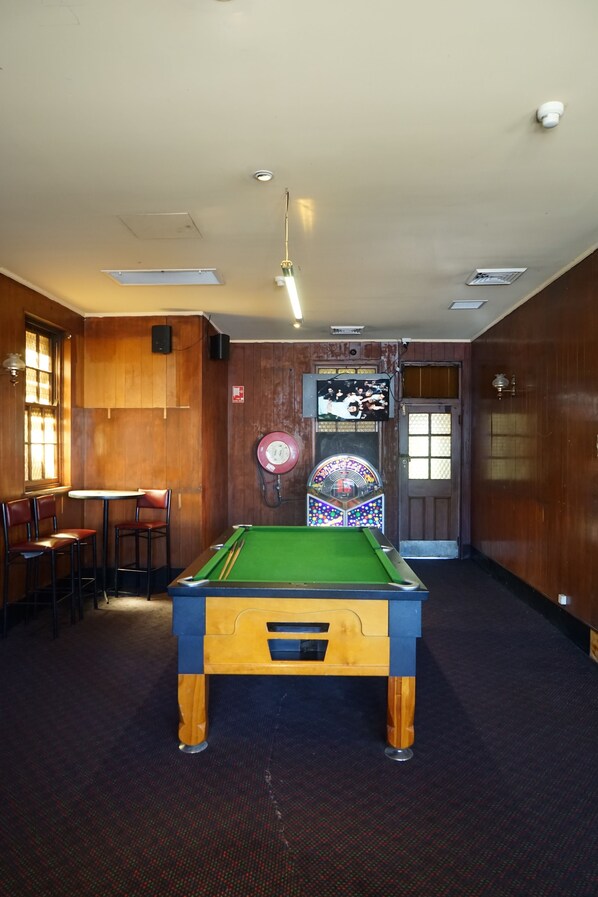 A pool table in a room with wooden walls, a bar area with stools, and a door with a window. - Rosehill Hotel (Granville)