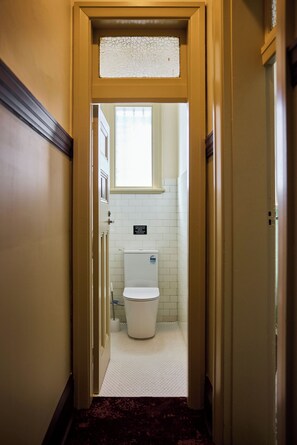 Un pasillo estrecho que conduce a un baño con un inodoro, un secamanos y un cartel que indica que las instalaciones son para todos los huéspedes.