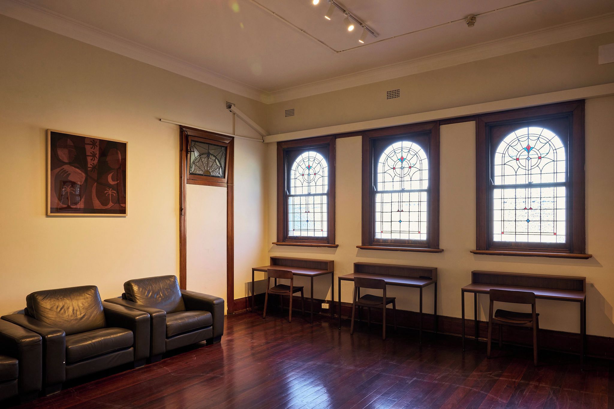 A room with a dark leather sofa, a wooden desk, and a chair. There are three large windows with stained glass.
