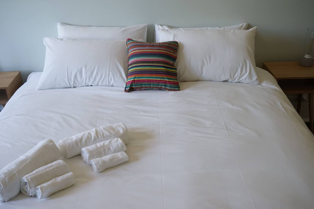 A neatly made bed with white linens, a striped pillow, and rolled towels on the side.