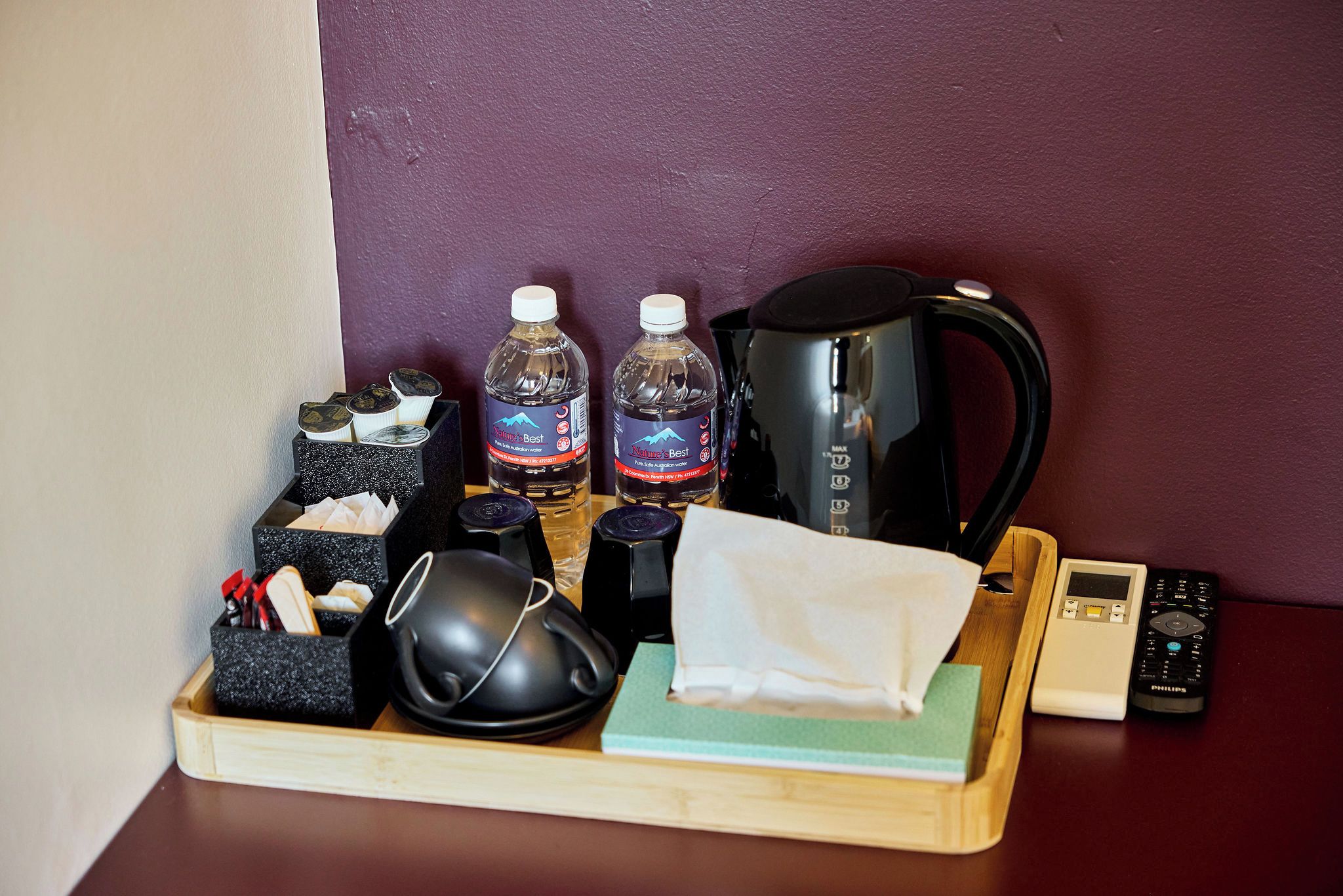A tray with a coffee maker, two bottles of water, a mug, a tissue, a remote control, and a box of tissues.