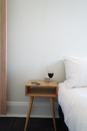 A small wooden bedside table with a smartphone, a glass of red wine, and a transparent object on a white bed in a hotel room. - Rosehill Hotel (Granville)