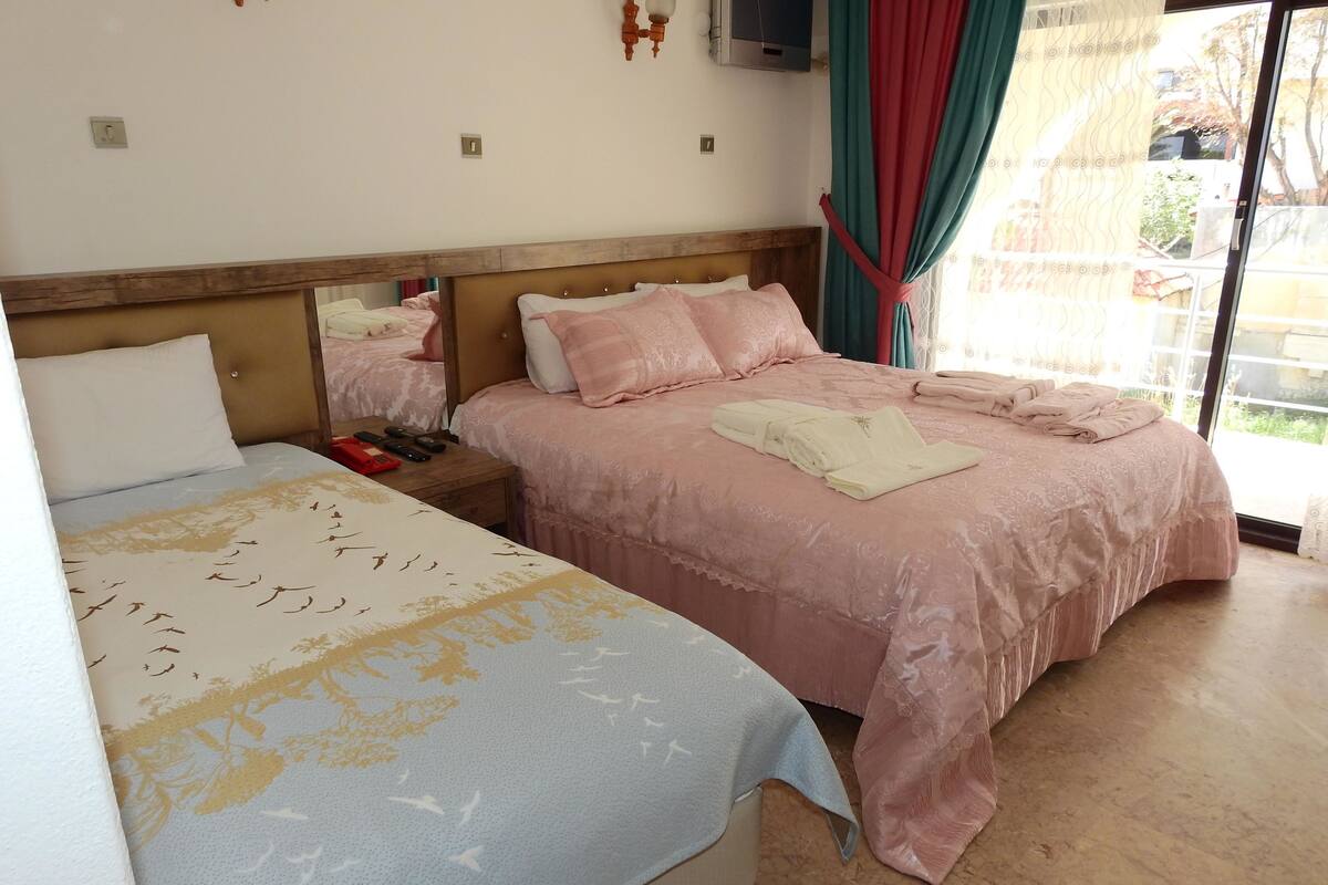 A hotel room with two beds, a wooden headboard, a small bedside table with a phone and remote controls, and a view of the outside through a glass door.