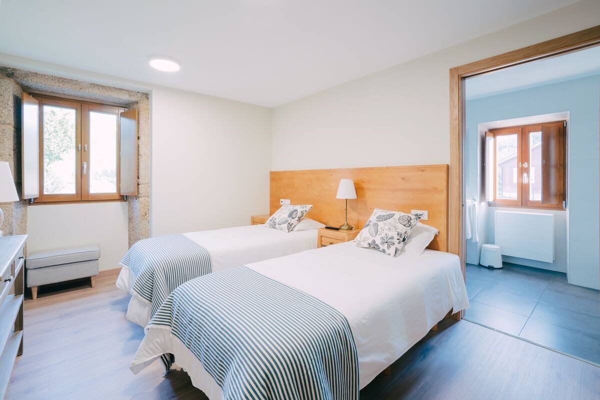 Quarto de hotel com duas camas, cabeceira de madeira, mesa de cabeceira com abajur, um banco e janela com vista.
