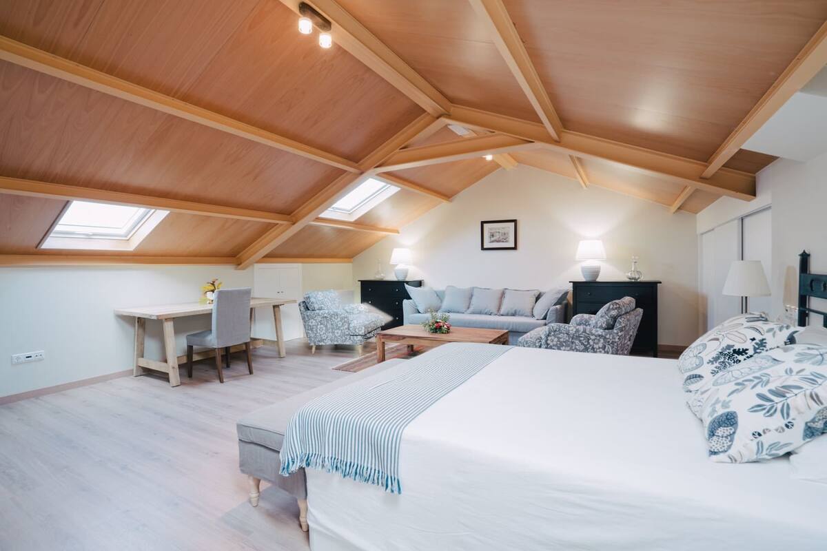 Um quarto amplo no estilo loft, com pé direito alto, claraboias, uma cama grande, uma escrivaninha e um sofá.