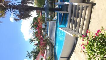 Zona de piscina con una piscina azul cristalina, rodeada de palmeras y flores de buganvilla rosadas.