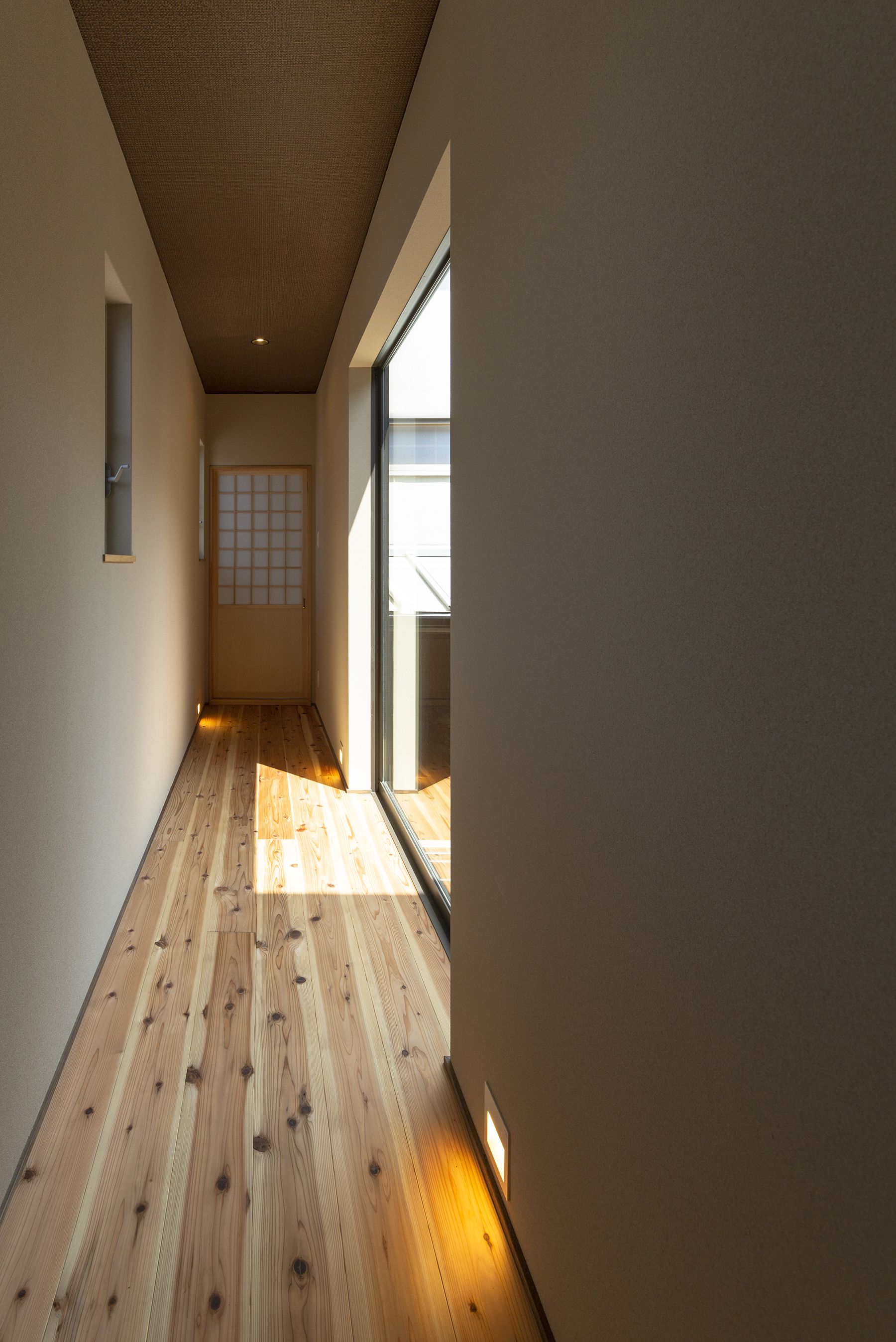 Un estrecho pasillo con piso de madera, una puerta corredera con diseño de cuadrícula y un ventanal luminoso a la derecha.