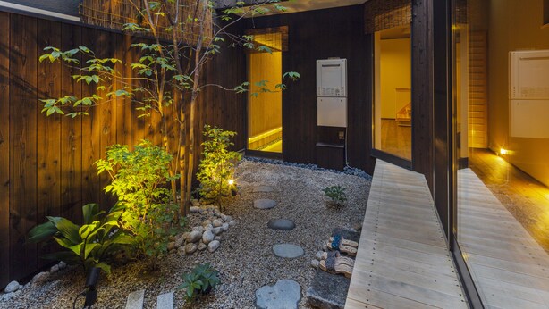 A modern building with a wooden exterior, a gravel pathway with stepping stones, and a small garden with plants and a tree. - hisashi (Kyoto)